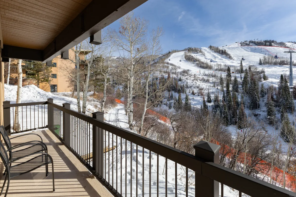 The view from a balcony looking out at snow-covered slopes, aspen trees, and mountain peaks. The balcony railing frames the scene, suggesting a private vantage point for enjoying sunsets or watching skiers glide by.