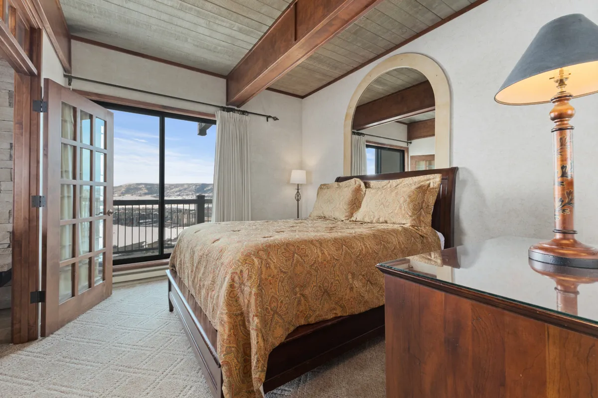 An inviting bedroom with ornate bedding and a large window offering scenic views of the mountains. A bedside lamp on a table adds to the tranquil and restful atmosphere, making it ideal for relaxation and quiet mornings.