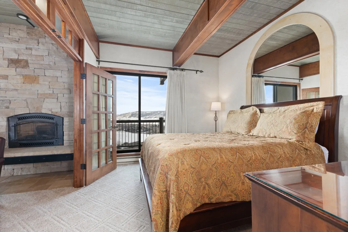 A tranquil bedroom featuring a bed with ornate bedding, a stone fireplace, and a large window framing mountain views. The space is inviting and evokes a sense of warmth and comfort, with natural light filtering in and a cozy ambiance enhanced by the fireplace.
