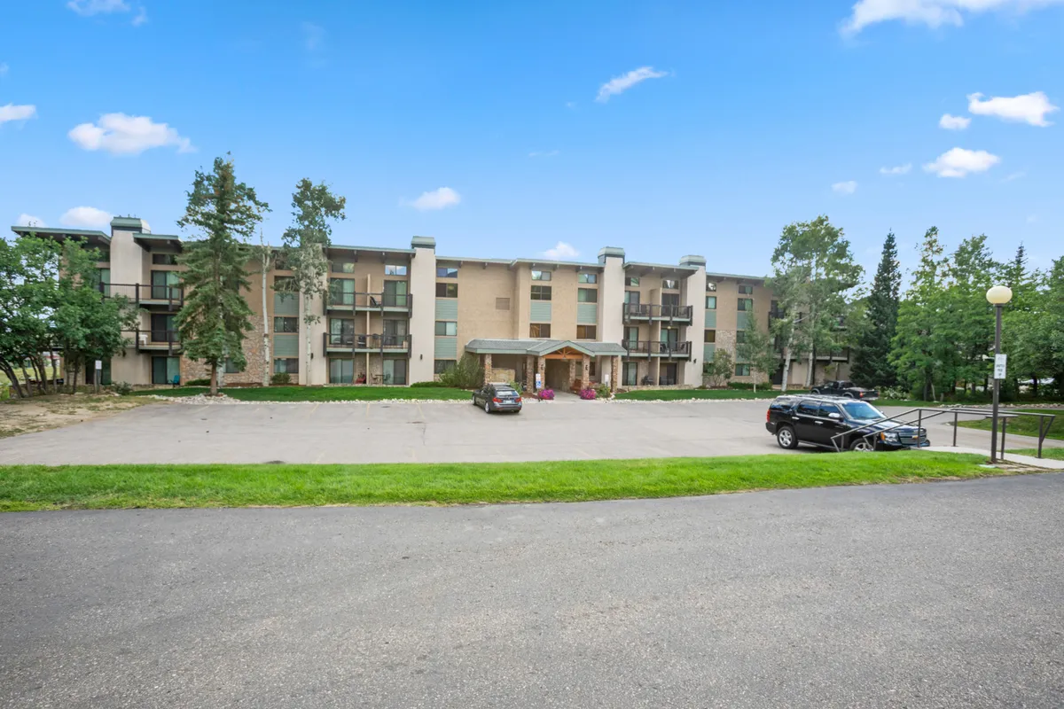 The photo shows the building's facade with an inviting entrance canopy and the parking area in the foreground. The pathway from the lot to the building is flat and easily navigable, lined with trees and surrounded by a welcoming atmosphere. The image provides a clear sense of how guests arrive from the parking lot to the main entrance, emphasizing the stress-free, step-free access.