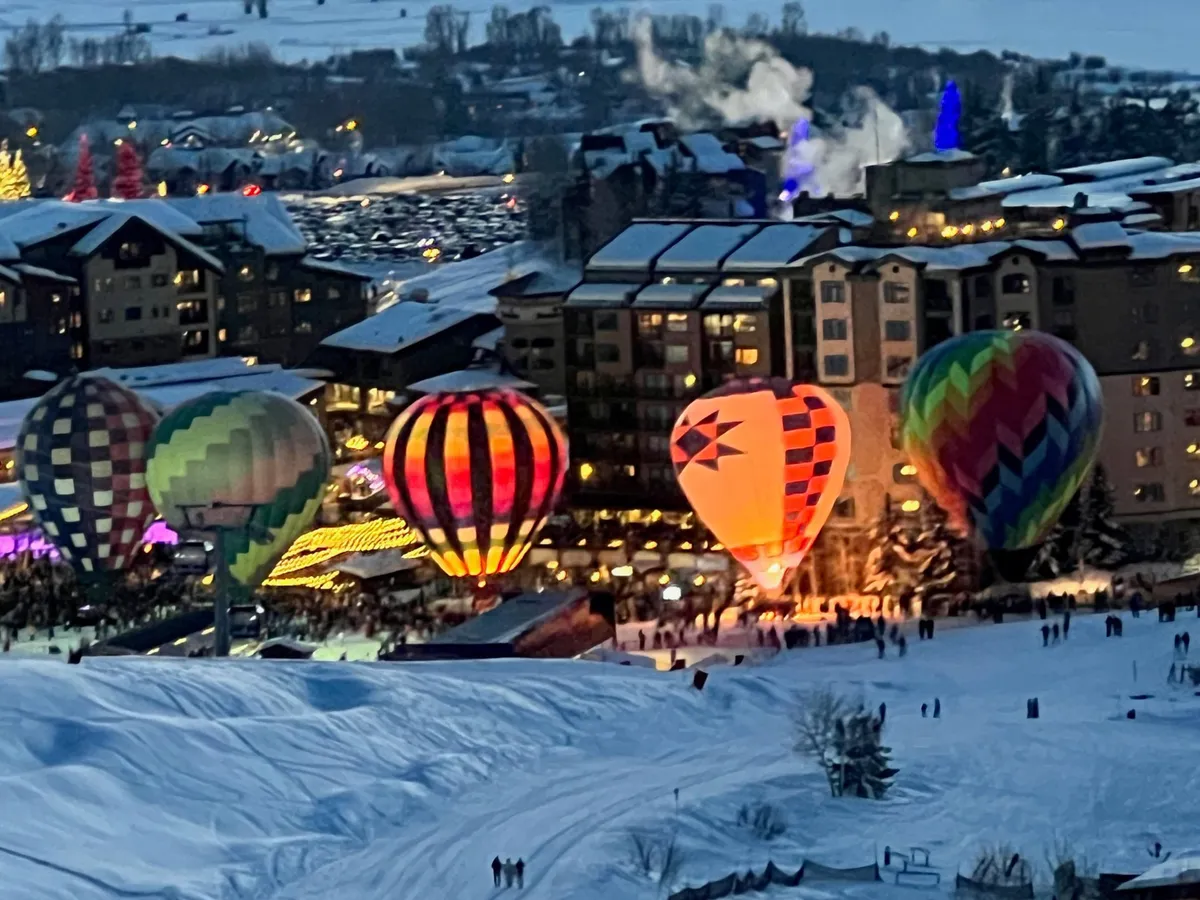 A festive outdoor scene featuring hot air balloons being inflated, illuminated buildings, and a crowd of people enjoying winter festivities. The joyful ambiance and the spectacle of the balloons highlight Steamboat’s celebrated events.