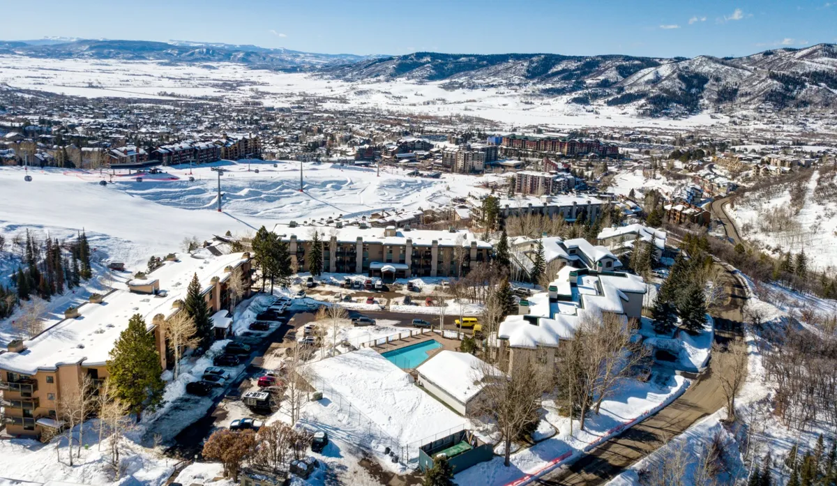 This exterior shot captures the residential building with the swimming pool in the foreground, snow-capped mountains in the background, and parking visible on the side. The pool area is an accessible amenity, with pathways leading to it, and the overall scene connects the condo to resort amenities and beautiful mountain scenery.