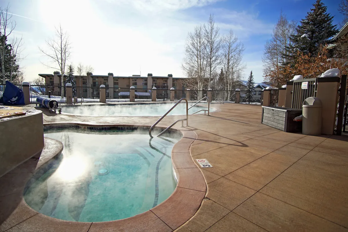 An inviting outdoor hot tub steaming in the cold air, surrounded by trees and a fenced area, set against the tranquil winter landscape. The setup suggests relaxation and socializing after a day spent outdoors.