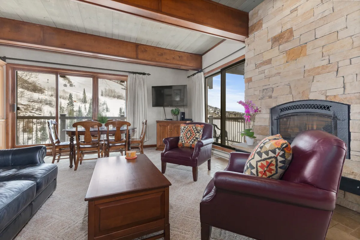 A spacious open-plan living area featuring a plush leather sofa, a large wooden dining table, and a stone fireplace. Expansive windows frame spectacular mountain views, flooding the space with natural light. The room is set up for family gatherings, cozy evenings by the fireplace, and game nights, with colorful throw pillows adding a welcoming touch.