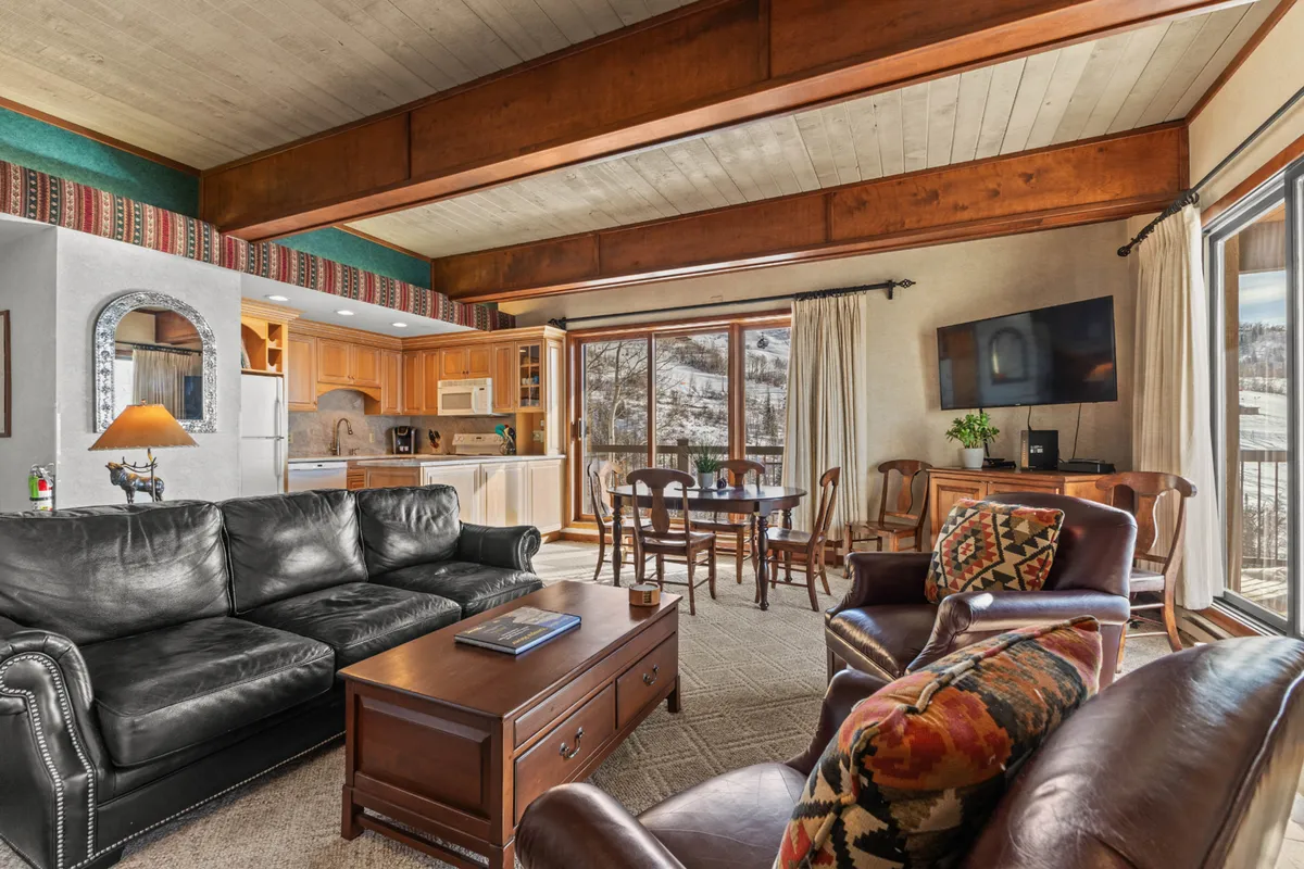 The inviting living and dining area showcases leather sofas, a wooden dining table, and large windows with snowy views. The kitchen area is visible in the background, emphasizing seamless flow between spaces. The scene reflects a warm, social atmosphere ideal for relaxing and sharing stories after a day's adventure.