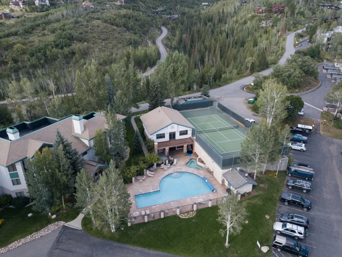 An exterior shot featuring the swimming pool, tennis court, surrounding greenery, and adjacent parking area. The image conveys an active, accessible environment with clear, level paths connecting key amenities and recreation areas.