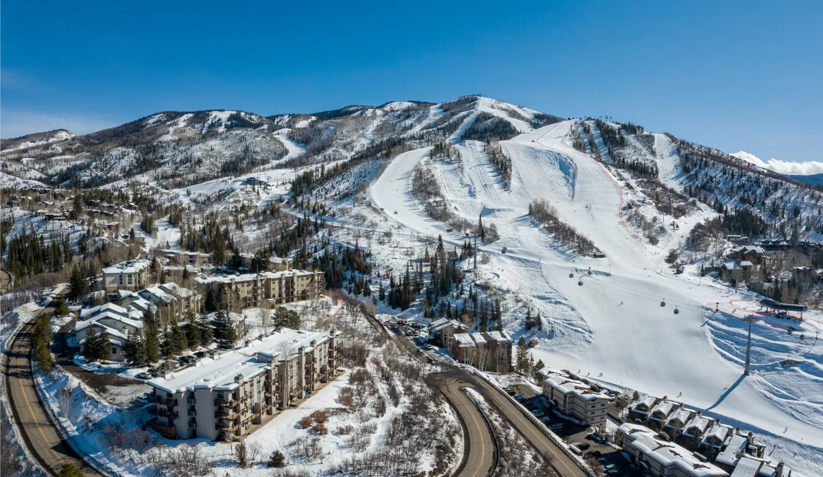 A sweeping view of snow-covered mountains and ski slopes, with multi-story resort buildings nestled among tall pine trees and gondola lifts visible overhead. The scene captures the heart of a vibrant ski resort area, where winter adventures await right outside the door.