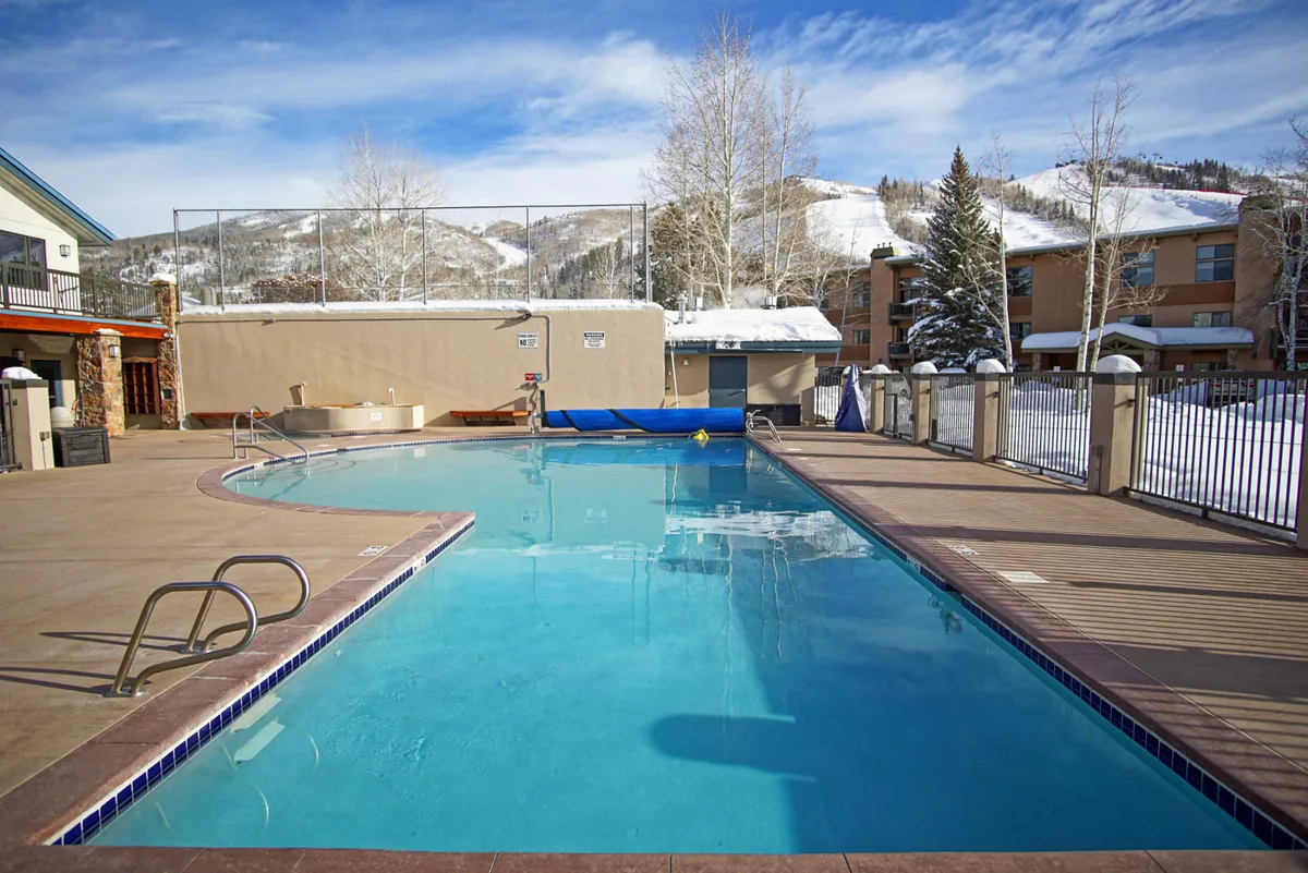 A serene pool area framed by snow-capped mountains and evergreen trees, with inviting lounge chairs and the condo building in view. The pool is set against a picturesque backdrop, suggesting relaxation and recreation opportunities beyond just snow sports.