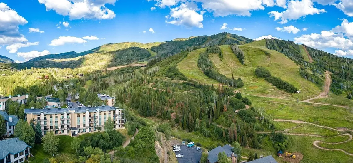 The hero image showcases the condo's exterior nestled among lush greenery, with sweeping views of the surrounding mountain slopes. At sunrise, golden light bathes the picturesque landscape, hinting at direct access to thrilling ski runs and tranquil nature trails. The buildings exude a cozy, welcoming vibe, perfectly situated for both adventure and relaxation.