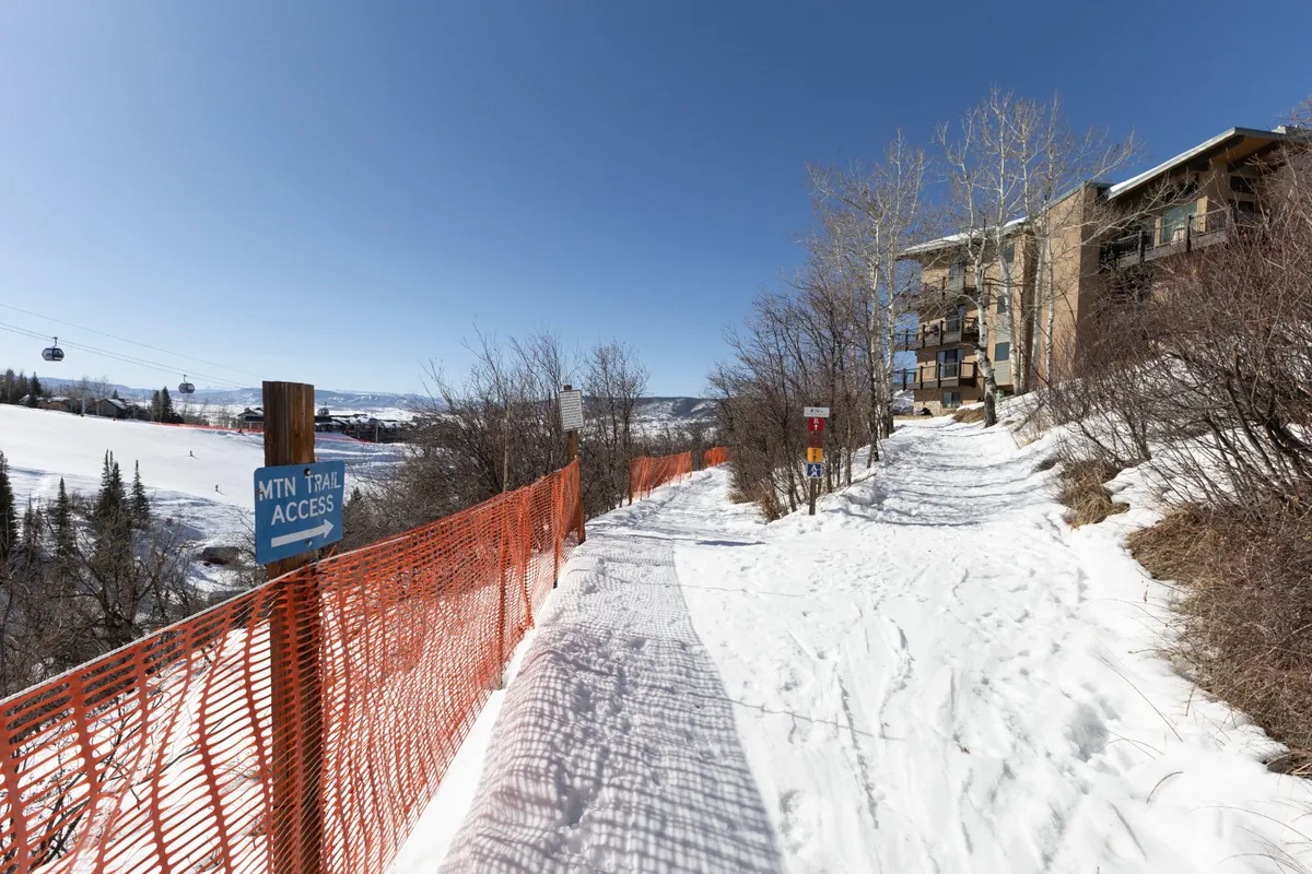 A snow-covered pathway leading to a mountain trail access sign, with a ski lift visible in the distance and a residential building nearby. The scene is inviting and hints at the versatile trail network accessible from the property, suitable for both skiing in winter and hiking or biking in warmer months.